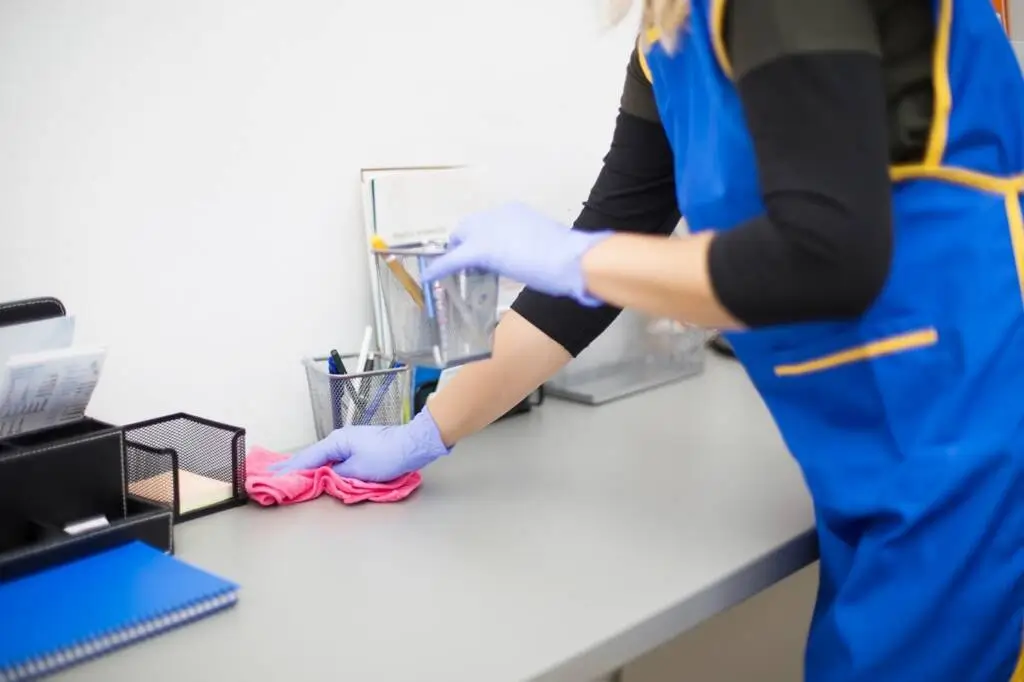 A professional day porter wearing rubber gloves holds a disinfectant spray bottle while cleaning and sanitizing an office table. - Paragon Peak Building Maintenance A professional day porter wearing rubber gloves holds a disinfectant spray bottle while cleaning and sanitizing an office table.