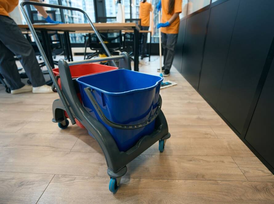 Janitorial cleaning service crew cleaning a coworking area floor, with a cleaning bucket in the foreground.