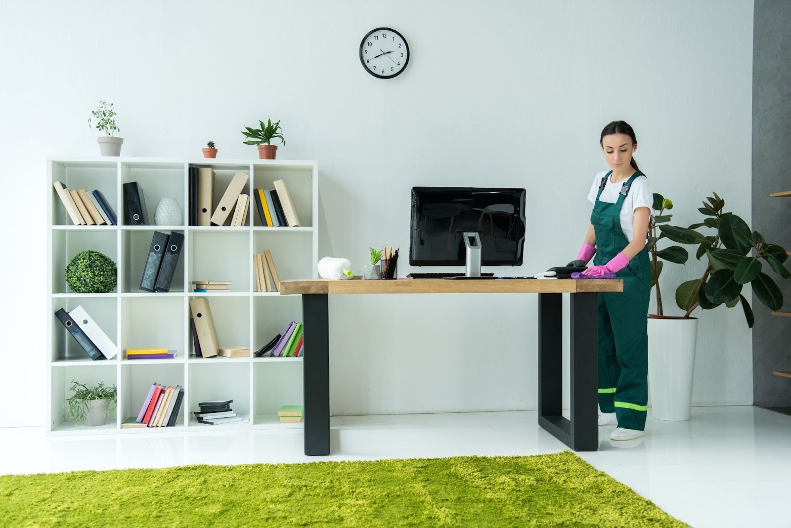 Female day porter in uniform and rubber gloves wipes a desk in a modern office during business hours