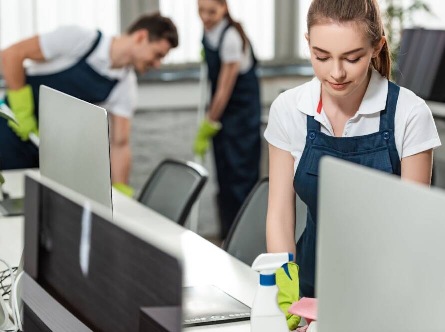 A female commercial cleaner wiping a desk while colleagues clean the office floor in the background