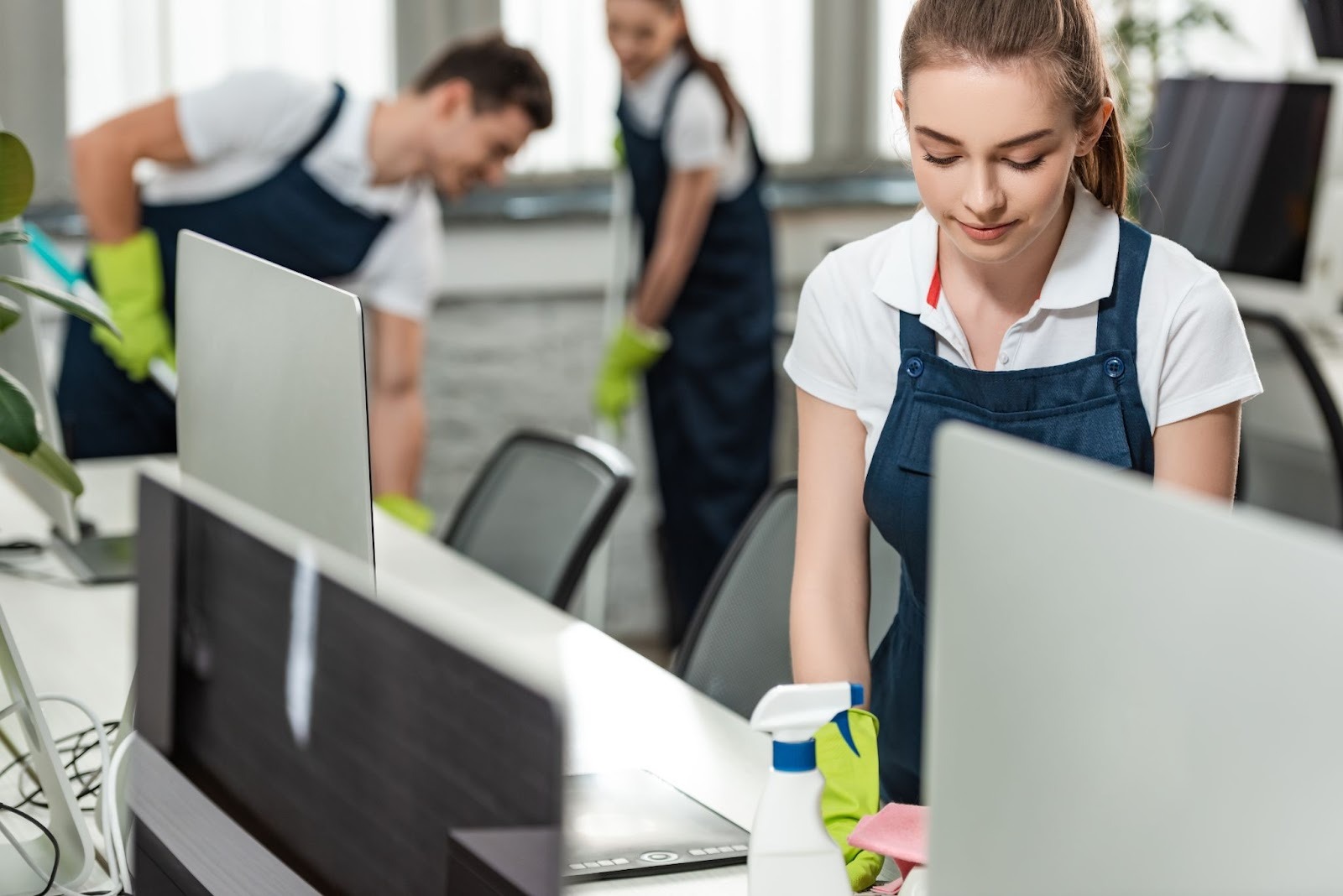 A female commercial cleaner wiping a desk while colleagues clean the office floor in the background