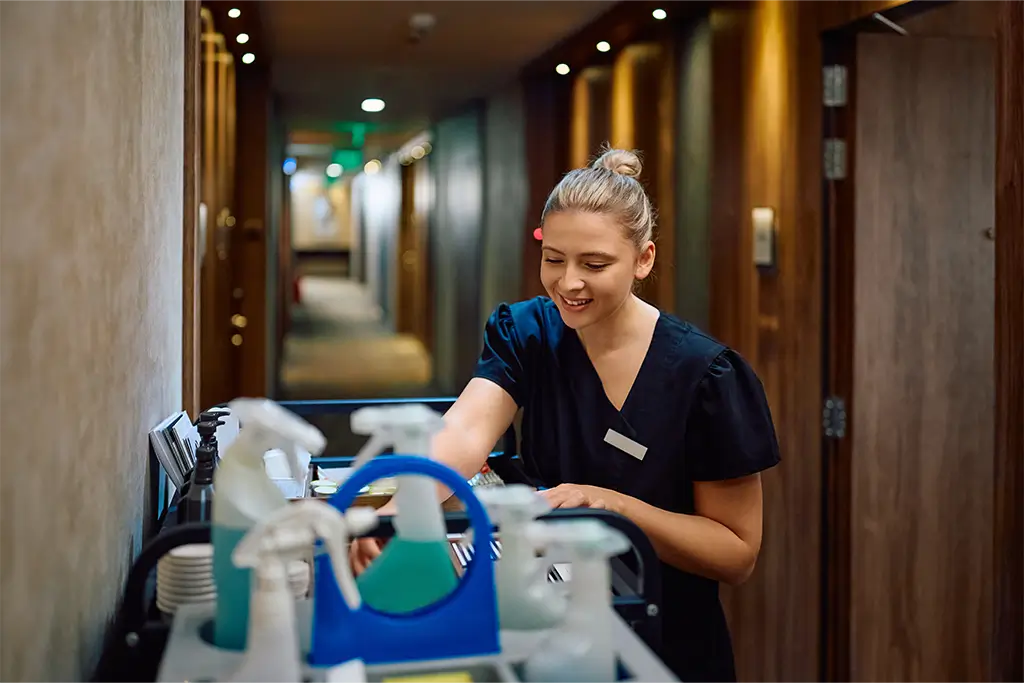 Cleaning lady reaching to grab her cleaning equipment in the middle of a hotel hallway. - Paragon Peak Building Maintenance Cleaning lady reaching to grab her cleaning equipment in the middle of a hotel hallway.